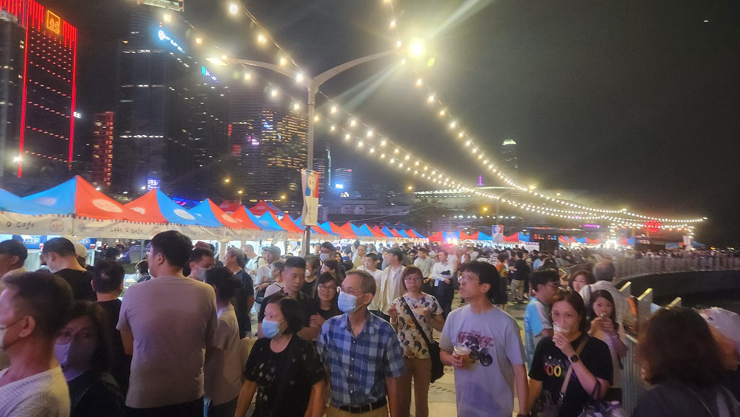 Crowds walking beneath string lights past red-and-blue market stalls at Waterfront Carnival